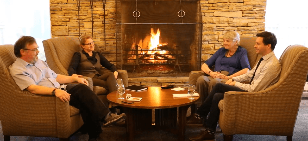 Four people sit in comfy chairs around a table with a lit fireplace in the background.