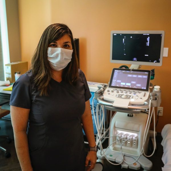A person with long brown hair, wearing a face mask and dark blue scrubs, stands in front of an ultrasound machine.